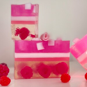 May include: Three pink and white layered soap bars with raspberry designs. The soap bars are arranged in a stack with the top bar slightly offset. There are several raspberries scattered around the soap bars.