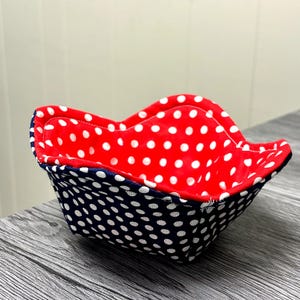 Puede incluir: Un cuenco de tela con interior rojo con lunares blancos. El exterior es azul marino con lunares blancos. El cuenco tiene un borde ondulado y festoneado. El cuenco está sobre una superficie de madera gris.