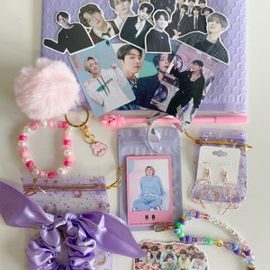 May include: A collection of purple accessories and BTS merchandise, including stickers, a pink pom-pom keychain, beaded bracelets, earrings, a scrunchie, and a pen. The items are arranged on a purple bubble mailer.