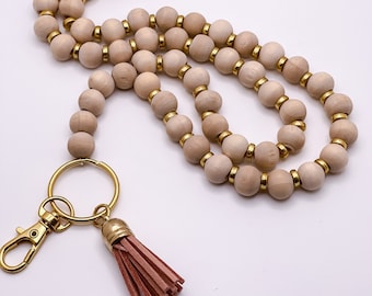 Może przedstawiać: Beżowy drewniany sznurek z koralikami, ze złotą metalową klamrą i różową frędzlą. Sznurek wykonany jest z okrągłych drewnianych koralików ze złotymi metalowymi akcentami.
