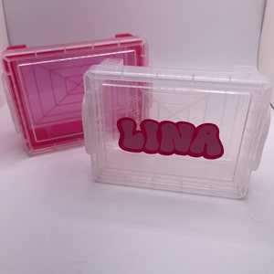 May include: Two clear plastic storage boxes. One is solid pink, the other is clear with the name "LINA" in pink bubble letters. Both boxes have a rectangular shape with a hinged lid and side handles.
