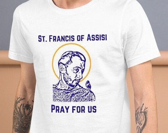 May include: White t-shirt featuring a blue and gold graphic of St. Francis of Assisi with the text "ST. FRANCIS OF ASSISI" above the image and "PRAY FOR US" below. The graphic is centered on the front of the shirt.