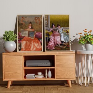 May include: Two framed paintings of women in vintage clothing, one reading a book and the other sitting with a friend. The paintings are hanging on a wall above a wooden entertainment center with a black media player and a white side table.