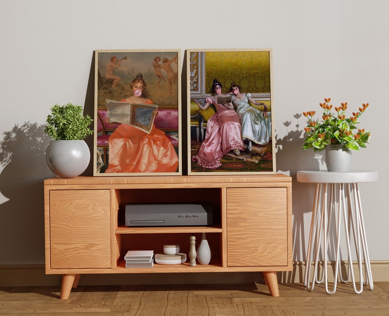 May include: Two framed paintings of women in vintage clothing, one reading a book and the other sitting with a friend. The paintings are hanging on a wall above a wooden entertainment center with a black media player and a white side table.