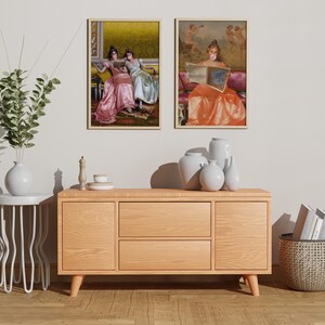 May include: Two framed paintings of women in vintage dresses, one reading a book and the other with a bubble, hanging on a white wall above a wooden dresser with two drawers, a white side table, and a wicker basket.