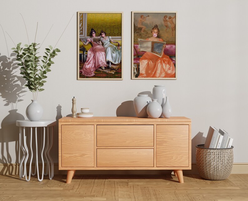 May include: Two framed paintings of women in vintage dresses, one reading a book and the other with a bubble, hanging on a white wall above a wooden dresser with two drawers, a white side table, and a wicker basket.