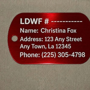 May include: A red metal tag with rounded corners and a hole for attachment. The tag features white text including a name, address, and phone number. The text reads: LDWF #, Name: Christina Fox, Address: 123 Any Street, Any Town, La 12345, Phone: (225) 305-4798.