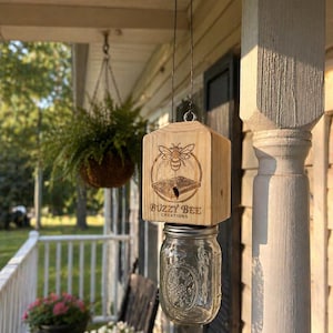 Könnte beinhalten: Ein hängender Bienenfutterspender aus Holz mit einem Glasbehälter. Auf dem Holz befindet sich ein eingraviertes Bienendesign und der Text "BUZZY BEE CREATIONS". Der Spender hängt an einem Draht. Im Hintergrund eine Veranda mit Pflanzen.
