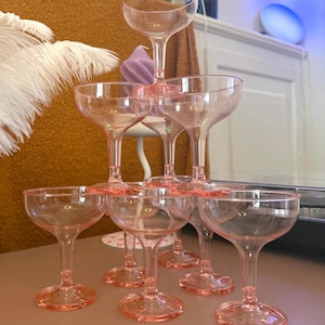 Puede incluir: Una pirámide de copas de champán rosas. Las copas están dispuestas en una formación escalonada, con una sola copa en la parte superior y números crecientes en los niveles inferiores. Las copas tienen una forma de copa clásica con un cuenco ancho y poco profundo y un tallo delgado.