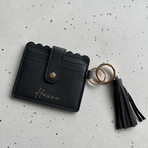 May include: Black leather cardholder with a scalloped edge and a gold-colored button closure. The cardholder has a gold-colored keychain with a black tassel. The name "Hanna" is written in gold script on the front.