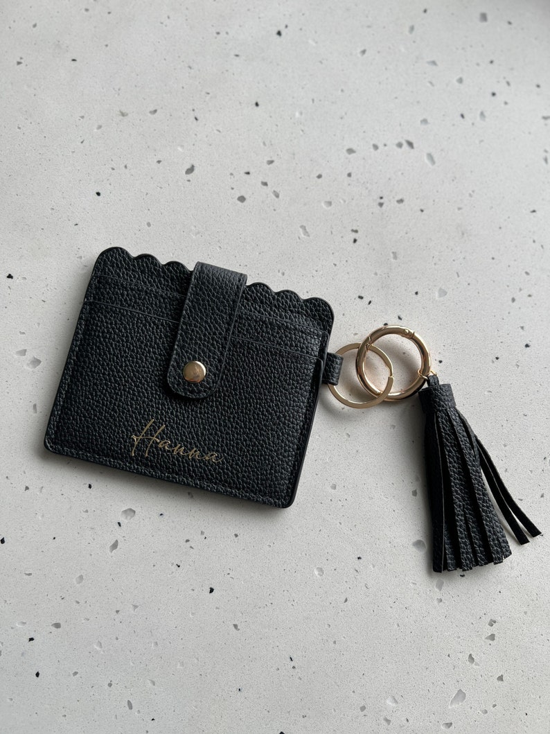 May include: Black leather cardholder with a scalloped edge and a gold-colored button closure. The cardholder has a gold-colored keychain with a black tassel. The name "Hanna" is written in gold script on the front.