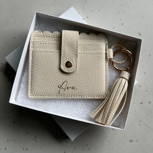 May include: Cream-colored cardholder with a scalloped edge, snap closure, and the name "Ava" printed on the front. Includes a matching tassel keychain with gold-tone hardware, presented in a white gift box.