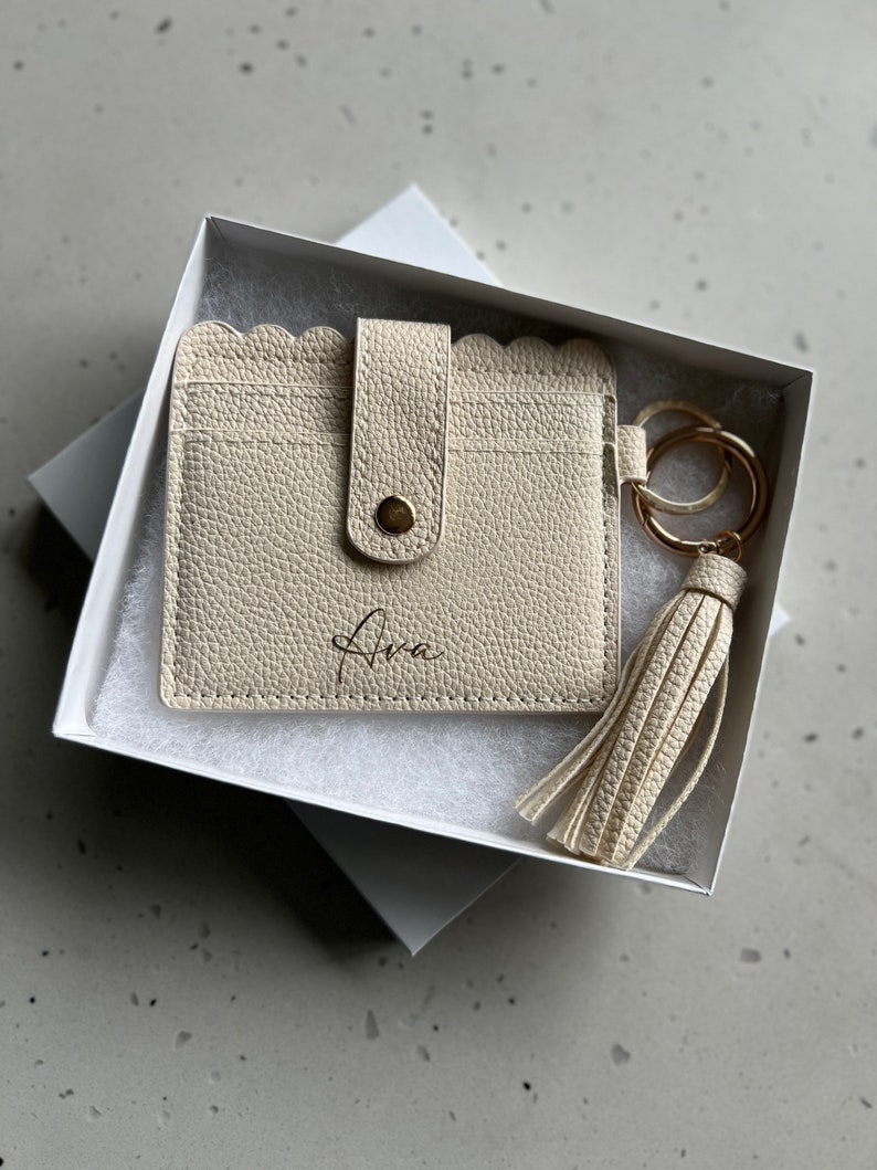 May include: Cream-colored cardholder with a scalloped edge, snap closure, and the name "Ava" printed on the front. Includes a matching tassel keychain with gold-tone hardware, presented in a white gift box.