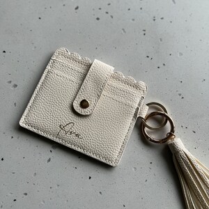 May include: Ivory-colored cardholder with a scalloped edge and a snap closure. The cardholder has a gold-tone keyring and a matching tassel. The name "Ava" is written on the front in a script font. The cardholder is made of textured material.