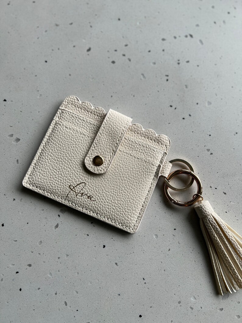 May include: Ivory-colored cardholder with a scalloped edge and a snap closure. The cardholder has a gold-tone keyring and a matching tassel. The name "Ava" is written on the front in a script font. The cardholder is made of textured material.