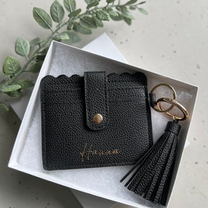 May include: Black leather cardholder with a scalloped edge, snap closure, and gold-tone hardware. Includes a matching tassel keychain and is presented in a white gift box. The name "Hanna" is printed in gold on the cardholder.