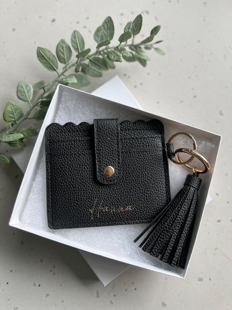 May include: Black leather cardholder with a scalloped edge, snap closure, and gold-tone hardware. Includes a matching tassel keychain and is presented in a white gift box. The name "Hanna" is printed in gold on the cardholder.