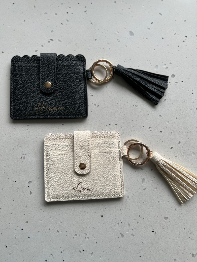 May include: Two cardholders with scalloped edges and keychains. One is black with "Hanna" in gold script, the other is off-white with "Ava" in gold script. Each has a gold button closure and a matching tassel.