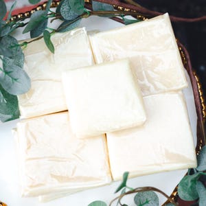 May include: A white, gold-rimmed serving tray holds several individually wrapped, square-shaped items. The items are a pale yellow color and are arranged on the white surface. Greenery with small leaves surrounds the tray.