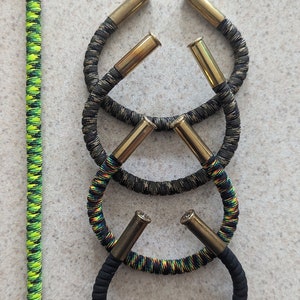 Puede incluir: Cuatro pulseras hechas con paracord y casquillos de latón. Las pulseras están apiladas una encima de la otra. Las dos pulseras superiores son de camuflaje negro y verde. Las dos pulseras inferiores son negras y multicolores. Una sola pulsera de paracord verde y negro está en el lado izquierdo de la imagen.