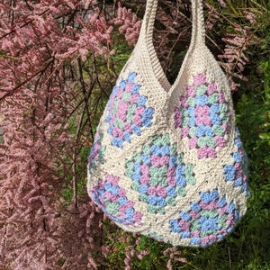 Puede incluir: Una bolsa de mano de crochet con un patr&oacute;n de granny squares en tonos de rosa, azul y verde. La bolsa est&aacute; colgada de una rama de &aacute;rbol con flores rosas.