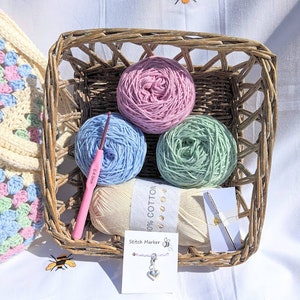 Puede incluir: Una cesta de mimbre llena de cuatro ovillos de lana en colores rosa, azul claro, verde y crema. La lana est&aacute; etiquetada como "100% algod&oacute;n". Tambi&eacute;n se incluye un gancho de ganchillo rosa y un marcador de punto.