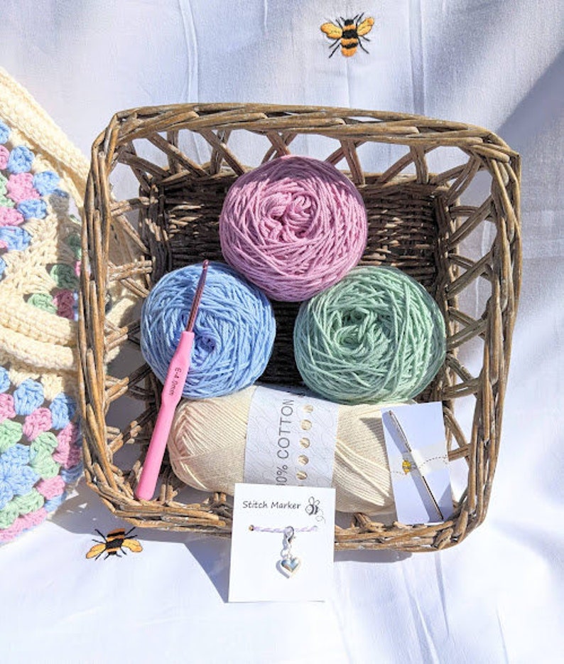 Puede incluir: Una cesta de mimbre llena de cuatro ovillos de lana en colores rosa, azul claro, verde y crema. La lana est&aacute; etiquetada como "100% algod&oacute;n". Tambi&eacute;n se incluye un gancho de ganchillo rosa y un marcador de punto.