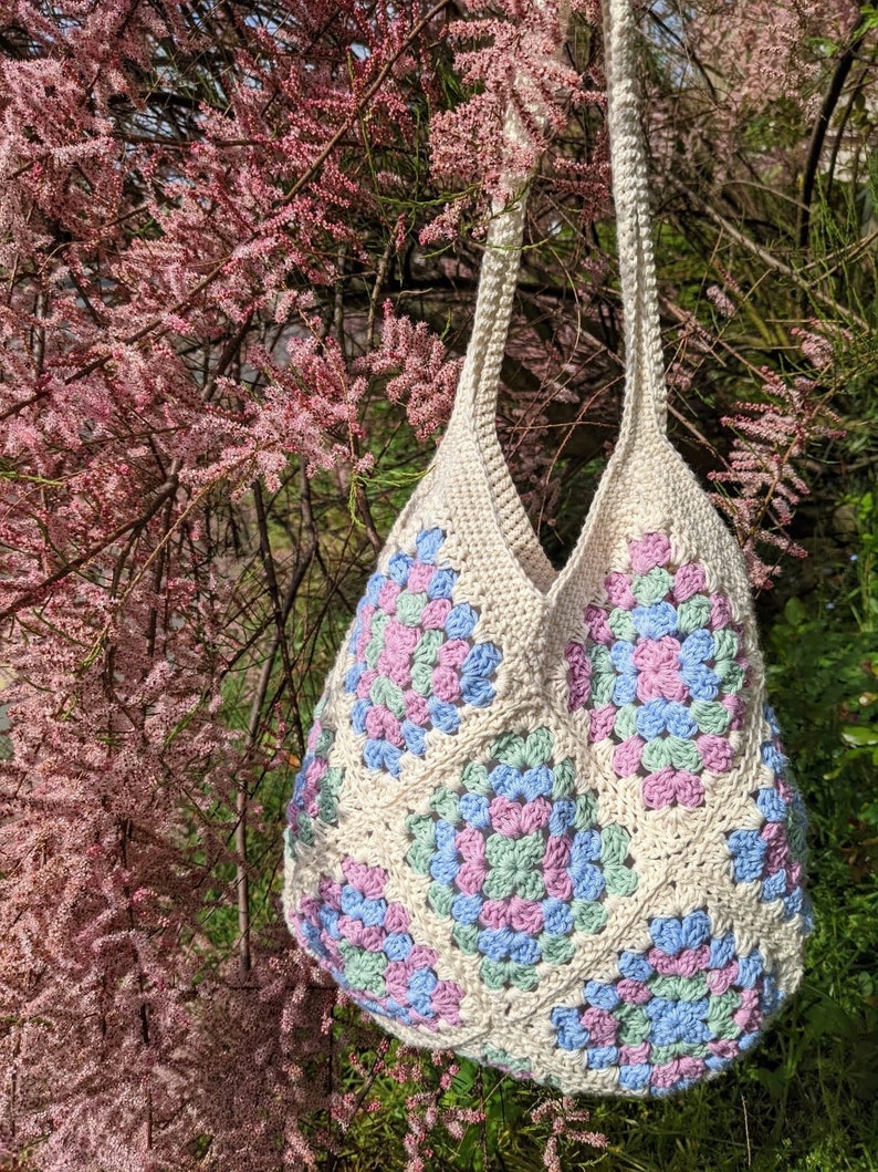 Puede incluir: Una bolsa de mano de crochet con un patr&oacute;n de granny squares en tonos de rosa, azul y verde. La bolsa est&aacute; colgada de una rama de &aacute;rbol con flores rosas.