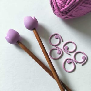 May include: Knitting supplies including two wooden needles with lavender tulip heads, lavender stitch markers, and a ball of lavender yarn. The items are arranged on a white surface.