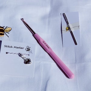 Puede incluir: Un ganchillo rosa con una etiqueta que indica 5,0 mm, un marcador de punto de plata con un dije en forma de coraz&oacute;n y una aguja de crochet de plata sobre un fondo blanco. La tarjeta del marcador de punto dice "Stitch Marker" con una ilustraci&oacute;n de abeja.