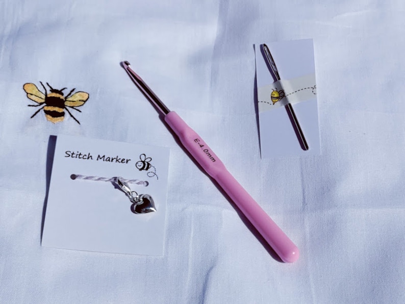 Puede incluir: Un ganchillo rosa con una etiqueta que indica 5,0 mm, un marcador de punto de plata con un dije en forma de coraz&oacute;n y una aguja de crochet de plata sobre un fondo blanco. La tarjeta del marcador de punto dice "Stitch Marker" con una ilustraci&oacute;n de abeja.