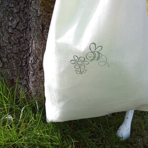 Puede incluir: Una bolsa de lona blanca con un dise&ntilde;o de abeja y flor en blanco y negro. La bolsa est&aacute; sobre un lecho de hierba verde.