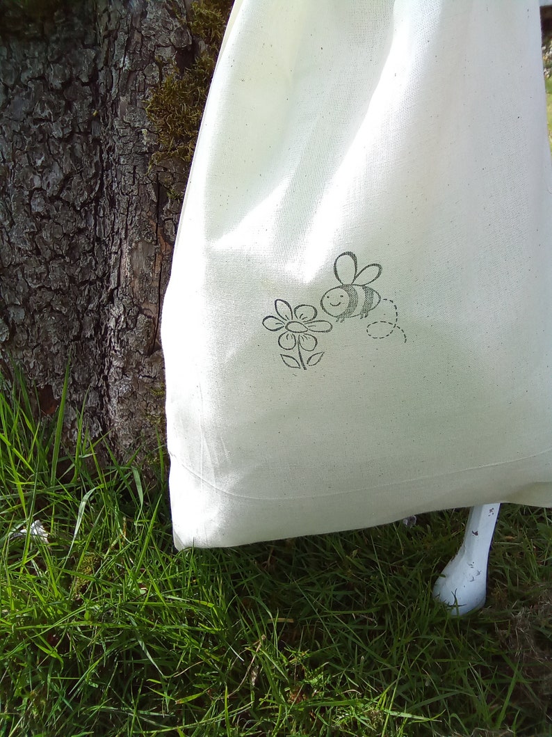 Puede incluir: Una bolsa de lona blanca con un dise&ntilde;o de abeja y flor en blanco y negro. La bolsa est&aacute; sobre un lecho de hierba verde.