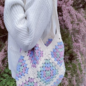 Puede incluir: Un bolso de mano blanco de crochet con un patr&oacute;n de granny squares en tonos de rosa, azul y verde. El bolso lo lleva una persona que lleva un su&eacute;ter blanco.