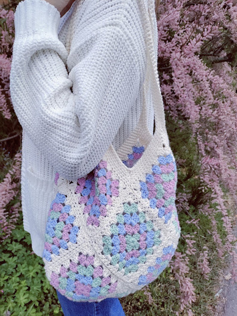 Puede incluir: Un bolso de mano blanco de crochet con un patr&oacute;n de granny squares en tonos de rosa, azul y verde. El bolso lo lleva una persona que lleva un su&eacute;ter blanco.
