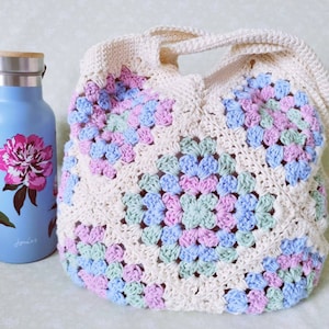 Puede incluir: Una bolsa de mano de crochet con fondo blanco y cuadrados de color rosa pastel, azul y verde. La bolsa tiene dos asas largas.