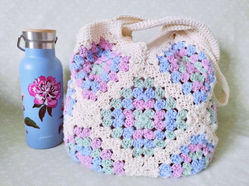 Puede incluir: Una bolsa de mano de crochet con fondo blanco y cuadrados de color rosa pastel, azul y verde. La bolsa tiene dos asas largas.