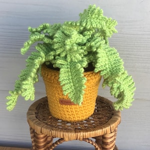 May include: A crocheted light green fern plant with textured, frond-like leaves potted in a mustard yellow crocheted container. The container features a small, rectangular brown leather-like tag with the word Handmade embossed on the front.