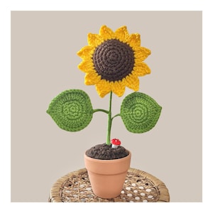 May include: A crocheted sunflower in a terracotta pot. The flower has yellow petals and a brown centre. Green leaves and stem support the flower. A small red and white mushroom sits in the soil. The pot is on a woven surface.