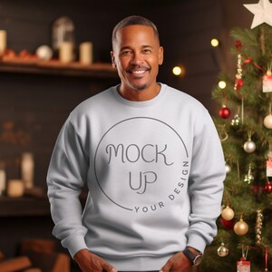 Puede incluir: Una sudadera blanca con un círculo gris que dice "Mock Up Your Design". La persona que lleva la sudadera está sonriendo y está de pie frente a un árbol de Navidad.