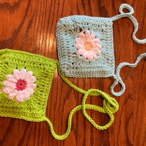 May include: Two crocheted flower squares with green and blue yarn. Each square has a white flower with a pink center. The squares are connected by a green yarn strap.