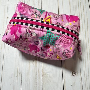 May include: A pink quilted makeup bag with a zipper closure. The bag is decorated with a colorful print of hearts, stars, lips, roller skates, and other girly designs.