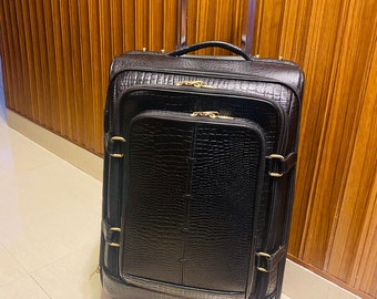 Op de afbeelding: Zwarte rolkoffer met een textuur die op krokodillenleer lijkt. De koffer heeft een telescopische handgreep, een handvat aan de bovenkant en goudkleurige hardware. De bagage staat op een betegelde vloer voor een houten wand.