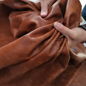 May include: Close-up of a large piece of brown leather. The leather is held up by two hands, showing its texture and flexibility. The leather has a rich, warm brown colour with variations in tone.