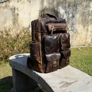 Könnte beinhalten: Ein großer, brauner Lederrucksack mit mehreren Außentaschen und einem oberen Griff. Die Tasche hat eine rechteckige Form mit einem Used-Look, der einen Vintage- oder robusten Stil suggeriert. Er steht auf einer Betonbank.