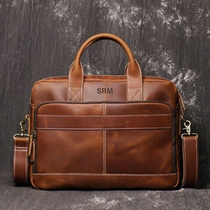 May include: A brown leather briefcase with a top handle and a detachable shoulder strap. The bag features a front pocket, visible stitching, and the initials "SRM" embossed on the front. The bag is approximately 16 inches wide.