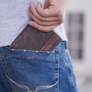 K&ouml;nnte beinhalten: Ein braunes Lederportemonnaie mit einem helleren braunen Akzent ist teilweise in einer blauen Jeans-Tasche zu sehen. Eine Hand greift in die Tasche, um das Portemonnaie herauszuziehen. Das Portemonnaie scheint neu und unbenutzt zu sein.