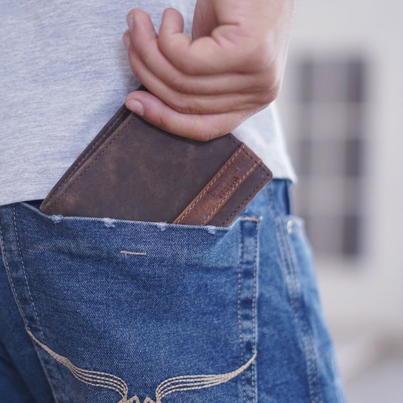 K&ouml;nnte beinhalten: Ein braunes Lederportemonnaie mit einem helleren braunen Akzent ist teilweise in einer blauen Jeans-Tasche zu sehen. Eine Hand greift in die Tasche, um das Portemonnaie herauszuziehen. Das Portemonnaie scheint neu und unbenutzt zu sein.