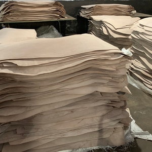 May include: Stacks of light brown wood veneer sheets, varying in size and shape, are piled in a warehouse setting. The sheets have a natural wood grain and are stacked on top of each other. Some stacks are on shelves.