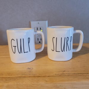 May include: Two white ceramic mugs with a slightly irregular, rounded square shape. The left mug features the word GULP in black, handwritten-style lettering, while the right mug features the word SLURP in the same black, handwritten-style font.